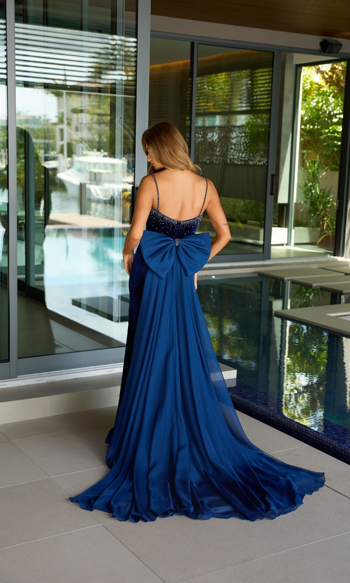 A woman in the Ava Presley 26522 long prom dress, featuring a large back bow, stands by a glass door near an indoor pool, facing away from the camera.